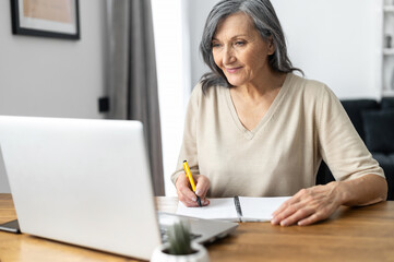 Charming mature mid-aged woman watching online webinars on the laptop sitting at the table at home, taking notes, e-learning. Senior business lady researching, analyzing tasks works in the home office