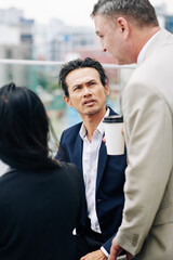 Frowning mixed-race businessman looking at talking mature colleague with cup of take away coffee
