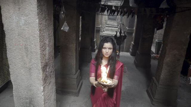 beautiful Indian girl in old stepwell wearing traditional Indian red saree, gold jewellery and bangles holding fire plate. Maa Durga shoot concept. Indian girl dancer in the posture of Indian dance
