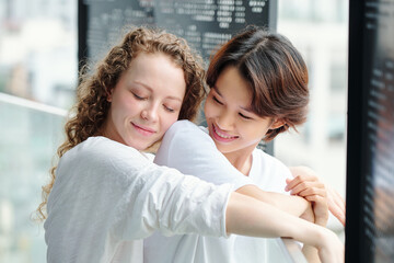 Happy pretty young curly woman hugging her girlfriend from behind when they are standing on balcony