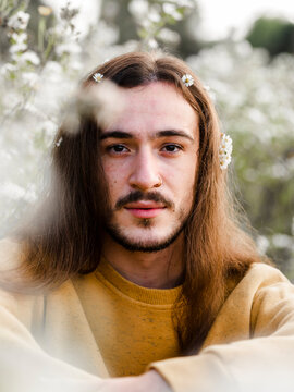 Romantic young long haired male in casual outfit sitting amidst blooming wildflowers and looking at camera while resting in summer countryside
