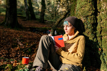 Peaceful female traveler leaning on tree trunk and writing in diary while relaxing in woods during summer journey