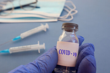 A gloved hand holds a flu vaccine against a background of masks and syringes, close-up