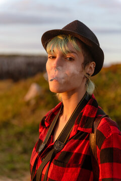 Carefree female hipster in hat and checkered shirt standing in nature and smoking cigarette at sunset