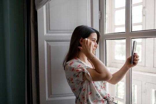 Side view of calm female with smartphone standing near window and taking picture of street through glass