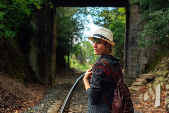 Back view of female traveler with backpack standing on rails in forest and looking at camera over shoulder during summer adventure