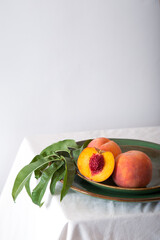 Peach in halves with bone in minimalistic style. Peaches with leaves on color green plate on table with white tablecloth. Ripe juicy peaches Still life. Harvest of peaches for food.