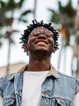 Low angle of delighted young African American male with dreadlocks wearing trendy denim jacket and eyeglasses smiling happily while standing on street