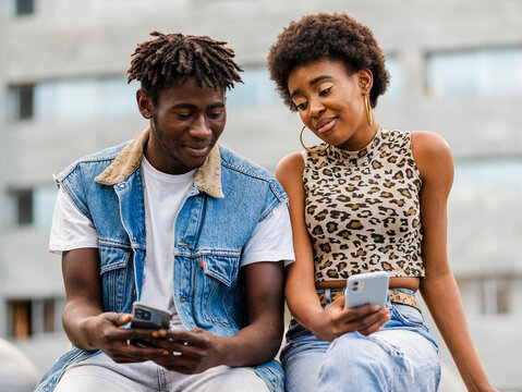 Cheerful young African American man and woman with curly hair dressed in trendy ripped jeans sitting on fence browsing on smartphone while spending time together in city
