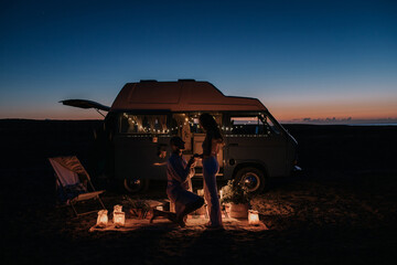 Side view of unrecognizable man making proposal for loving woman while standing on knee near illuminated van at night