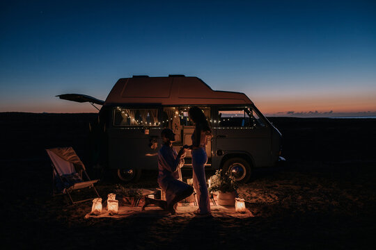 Side view of unrecognizable man making proposal for loving woman while standing on knee near illuminated van at night