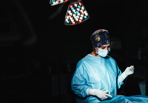 Side view of focused professional female surgeon with surgical tools and thread finishing operation of patient in operating theater