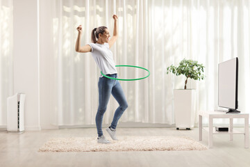 Young woman spinning hula hoop at home in a living room - obrazy, fototapety, plakaty