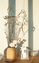 Wicker vase with dry branches and a figurine of a house on the window in the rays of the sun