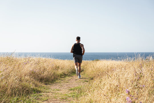 Muscular male jogger running along path in meadow during training on background of sea in summer