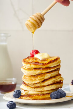 Pouring honey on a stack of fluffy breakfast pancakes with berries and butter