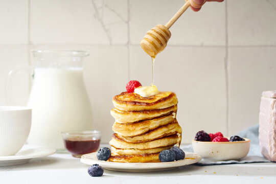 Pouring honey on a stack of fluffy breakfast pancakes with berries and butter