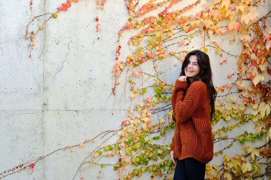 Cheerful young long haired brunette in warm knitted sweater standing near stone wall with colorful autumnal foliage and looking at camera