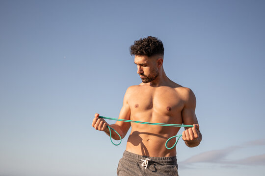 Muscular male bodybuilder with naked torso standing on seashore and doing exercises with resistance band during workout in summer