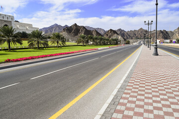 Beautiful Al Saidiya Street lined by lampposts, a green grass, flowers, palm trees and mountains. Muscat, Oman.