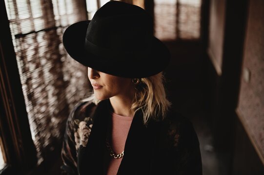 Charming pensive young female in elegant vintage clothes and hat standing in front of window in dark room