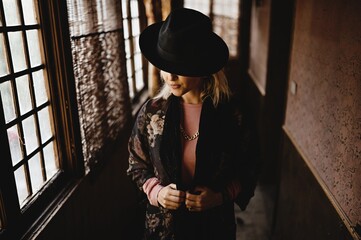 Stylish female in elegant vintage outfit and hat standing in narrow hallway of old house and looking at mirror