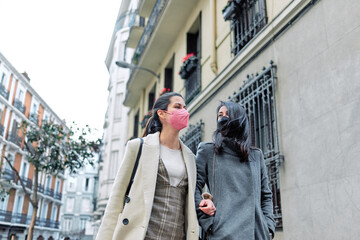 Two happy women talking and walking in the street with their face masks on.