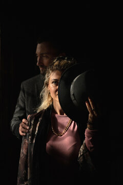 Adult Woman Covering Face With Hat And Looking At Camera While Standing With Beloved Man Near Window In Dark Room Of Old House