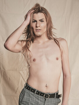 Androgynous young shirtless guy with long hair looking at camera against beige background
