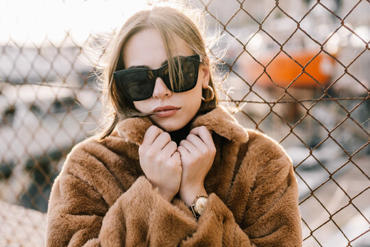 Young determined female in sunglasses and warm jacket standing near net fence and looking at camera