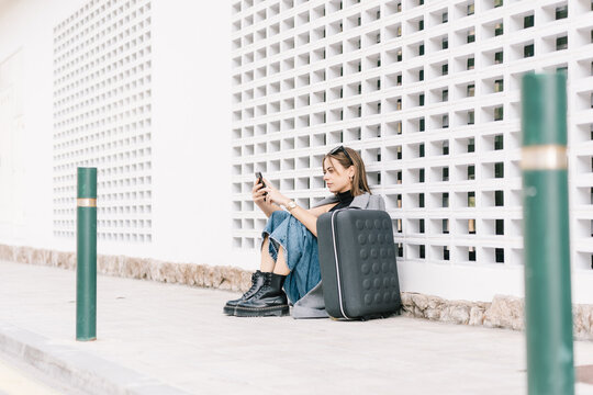 Side view of female tourist with luggage sitting alone on pavement and browsing mobile phone while getting lost in city