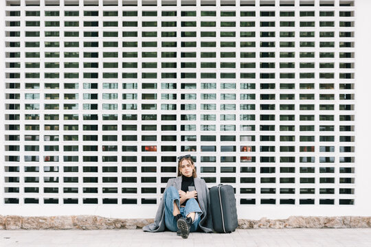 Female tourist with luggage sitting alone on pavement while getting lost in city looking at camera