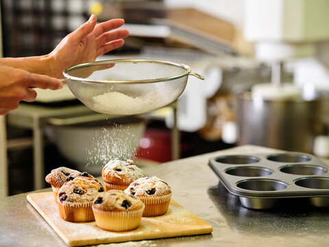 Side view of crop unrecognizable confectioner with sieve sprinkling sugar powder over freshly baked muffins with raisins while working in professional kitchen