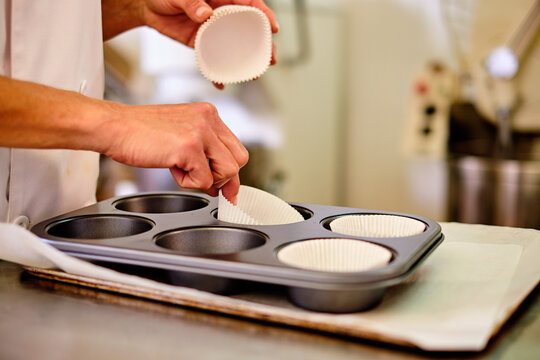 Side view of crop anonymous confectioner arranging paper cups into metal muffin tray before filling with dough while preparing pastry in kitchen