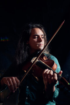 Talented male musician playing wooden violin with closed eyes while sitting in dark room