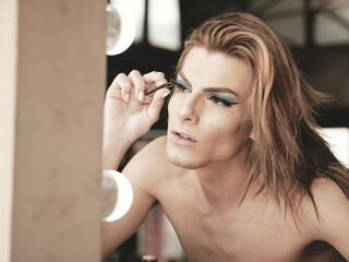 Young androgynous male model with manicure using tweezers to apply fake eyelashes in front of mirror in studio