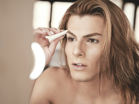 Young androgynous man with long hair applying foundation on face with cotton pad in front of mirror in studio