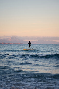Male surfer in wetsuit rowing on paddle board on sea water on background of mountains at sunset