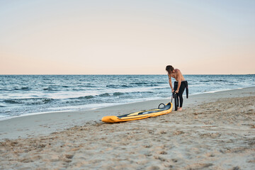 Man in wetsuit inflating paddle board at the seashore on sandy beach