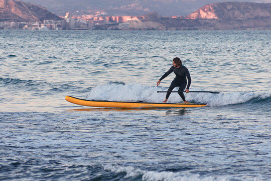 Male surfer in wetsuit rowing on paddle board on sea water on background of mountains at sunset