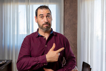 Young handsome bearded man wearing a maroon shirt pointing aside worried and nervous with his index finger