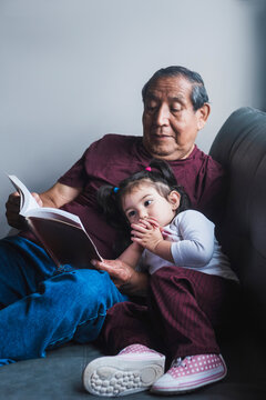 Senior ethnic grandfather reading interesting story in book for cute little girl while relaxing together on sofa at home