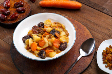 A dish of Jewish cuisine tsimes with carrots and dates in a white plate on a round stand next to a spoon carrots
