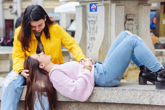 Gentle couple of lesbian females resting on stone bench on street while smiling and looking at each other