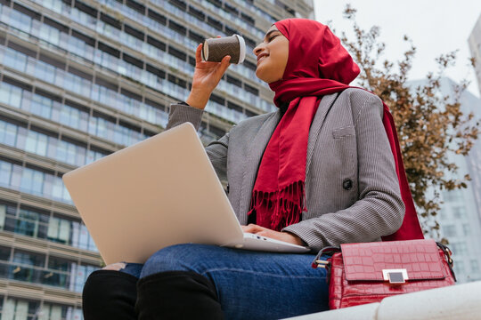 From below of Arab female entrepreneur in hijab sitting on street with laptop and dirking coffee to go while working on remote project in city