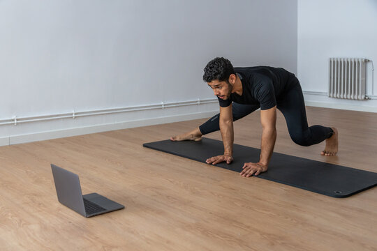 Concentrated ethnic male athlete practicing on mat during training while watching video tutorial on laptop