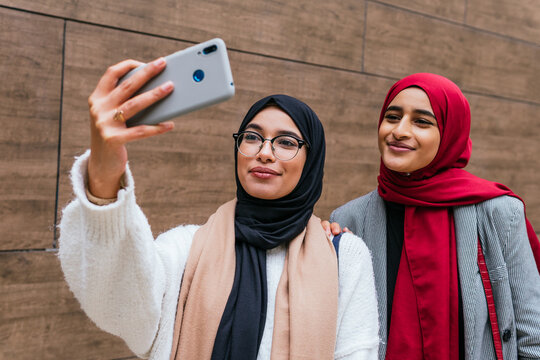 Low angle of Arab female best friends in hijab standing in street and taking selfie on smartphone