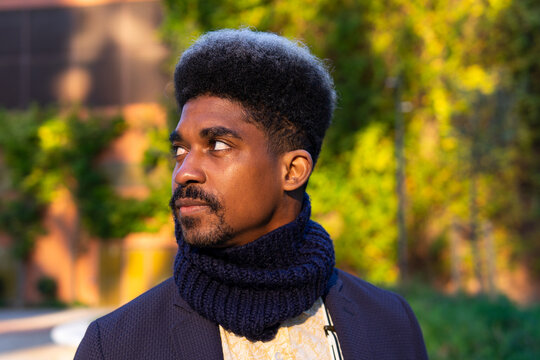 Pensive African American male with curly hair and in stylish outfit standing in urban park on sunny day and looking away