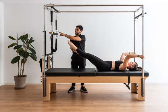 Slim female in sportswear doing exercises on cadillac reformer with help of professional trainer during pilates workout