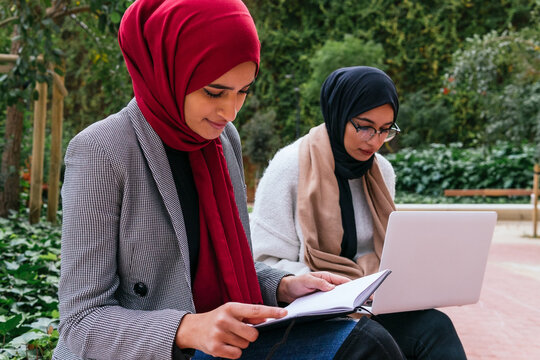 Content Arab female students in headscarf sitting on bench in green garden of campus and preparing for exams while reading notes and using laptop